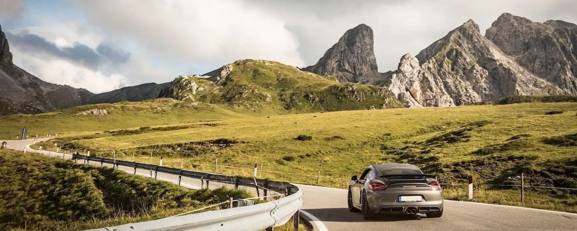 A Porsche Cayman drives over a mountain pass with a great view of the mountains.